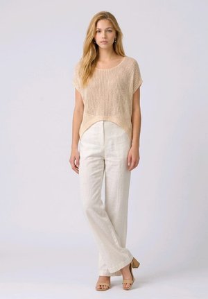 Mujer con cabello rubio largo lleva un top beige de punto de manga corta, pantalones blancos de cintura alta y tacones beige de punta abierta, de pie sobre un fondo claro.