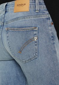 Jeans de mezclilla azul claro con una etiqueta de cuero marrón que dice "DONDUP". Detalles notables incluyen un diseño de costura único y un pequeño bolsillo decorativo.
