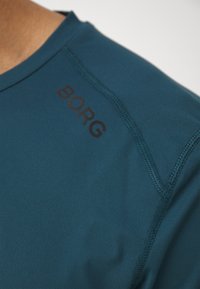 Tyrkysové sportovní tričko s výstřihem do V. Má černý text "BORG" na rameni, hladkou tkaninu a zesílené švy.