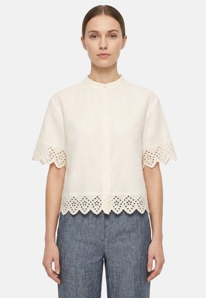 Femme portant un chemisier blanc à manches courtes avec des bords festonnés en dentelle ajourée et un pantalon gris taille haute, debout devant un fond uni.