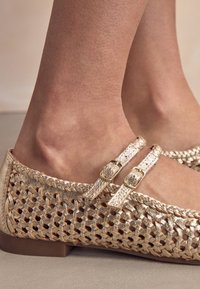 Lygūs, metalinio aukso spalvos batai su austu odos paviršiumi, turintys du reguliuojamus dirželius su auksinėmis sagtimis ir apvalia nosimi.