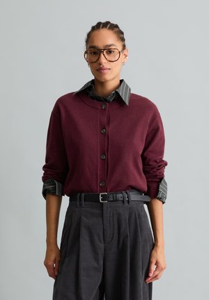 Femme aux cheveux tressés et grandes lunettes portant un pull bordeaux boutonné sur une chemise rayée et un pantalon plissé sombre taille haute.