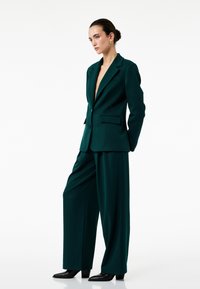 Mørkegrøn skræddersyet blazer med notcherede klapper, parret med vide bukser. Glat stof og elegant design. Sorte spidse hæle.
