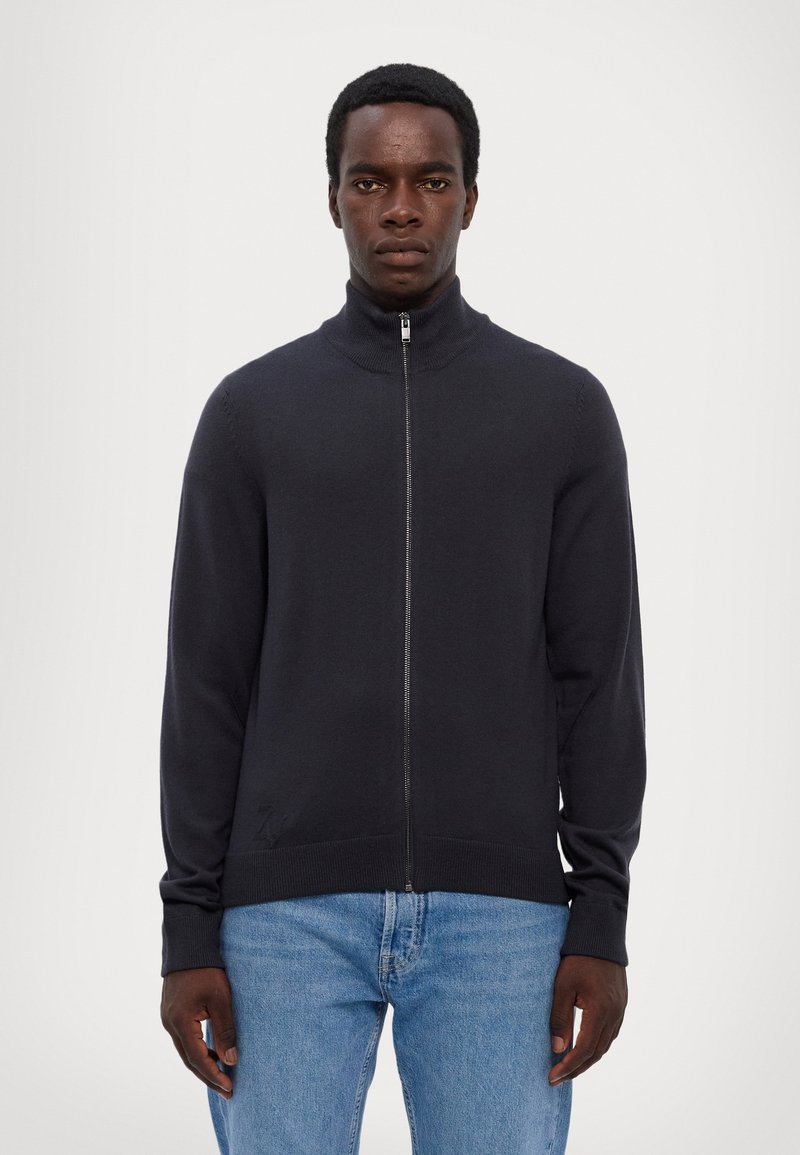 Dunkelblaues, mit Reißverschluss versehenes Cardigan mit hohem Kragen, gerippten Bündchen und Saum. Sanfte Strickstruktur. Aufgenähtes Logo nahe dem Saum. Über blauen Jeans getragen.