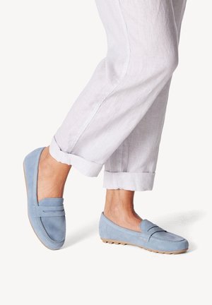 Benen in lichtgrijze manchetbroek en lichtblauwe suède loafers met rubberen zolen op een witte achtergrond.