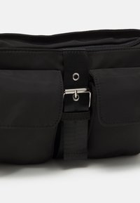Borsa nera in nylon con una zip superiore, tasche anteriori con pattina e una larga tracolla regolabile con fibbia in metallo e dettagli hardware.
