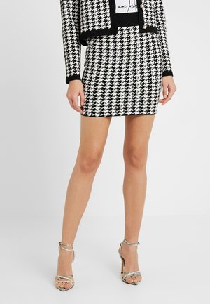Femme portant une mini-jupe pied-de-poule noire et blanche et une veste courte assortie avec un t-shirt graphique et des sandales métalliques à talons hauts.
