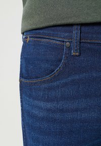 Jeans de mezclilla azul oscuro con una textura suave, que cuentan con un bolsillo delantero, costuras naranjas en contraste y un detalle de hardware de botón negro.