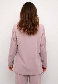 Blazer de corduroy rosa claro con mangas largas y corte recto, que presenta una textura de canalé vertical. Combinado con pantalones a juego.