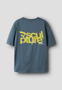 Pelēks kokvilnas T-krekls ar lielu dzeltenu grafisku tekstu aizmugurē, kas lasāms "skulptūras kolektīvs", un papildus mazāku tekstu.