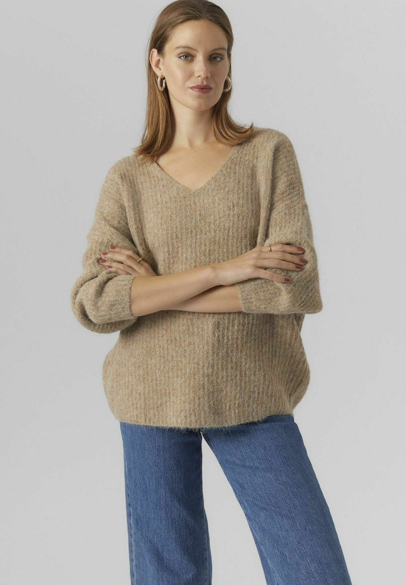 Beige stickad tröja med V-hals, sänkta axlar och strukturerat mönster. Kombineras med mörkblå jeans med en avslappnad passform.