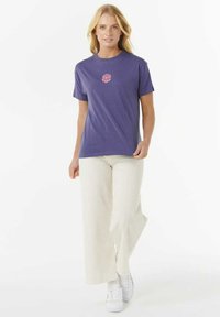 Lila T-Shirt mit einem kleinen, gemusterten pinken Design in der Mitte, kombiniert mit weit geschnittenen weißen Hosen und weißen Sneakers.