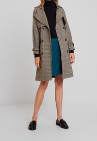 Trench coat a scacchi nei toni beige, nero e rosa, con un grande colletto, bottoni doppi, e polsini con cintura, abbinato a una gonna color teal.