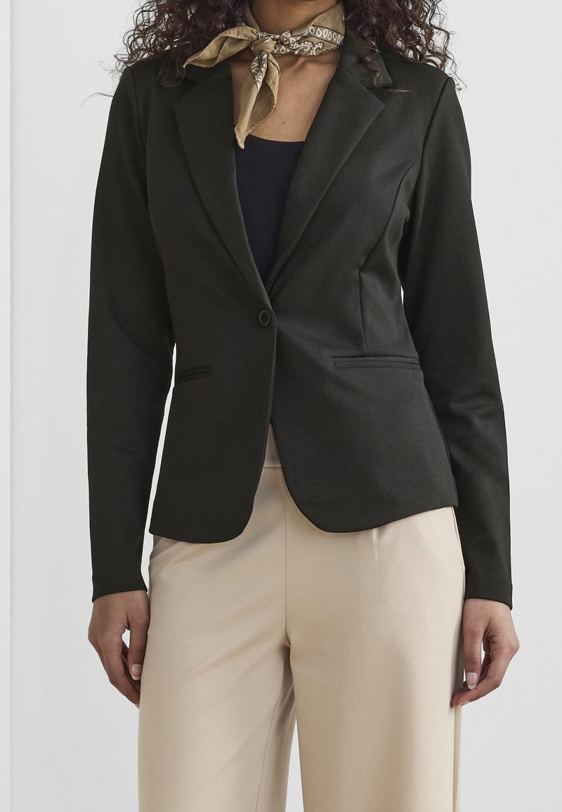 Femme portant un blazer noir cintré, un pantalon beige, un haut foncé et un foulard léger marron à motifs autour du cou sur un fond uni.