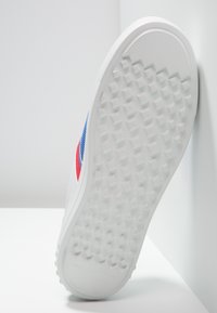 Semelle de chaussure de sport blanche avec un motif texturé antidérapant, comprenant des accents rouges et bleus sur le côté. Construction plate en caoutchouc durable pour une bonne adhérence.