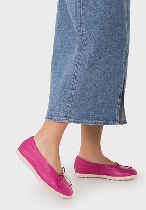 Persona que lleva pantalones cortos de mezclilla azul y bailarinas de cuero rosa brillante con detalle de lazada, de pie sobre un fondo claro.