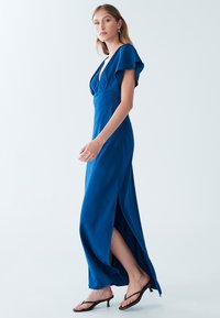 Dlhé modré šaty z hladkej látky s hlbokým výstrihom do V, nadýchanými rukávmi a vysokými rozparkami na oboch stranách, doplnené čiernymi sandálmi na opätku.