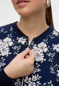 Navy floral bomber jacket with white flowers, zip front, and ribbed collar, featuring smooth fabric and subtle green leaf accents.