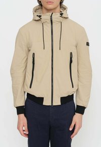 Giacca leggera beige con cappuccio, dotata di zip nere, coulisse e polsini a costine. Due tasche anteriori con zip, tessuto morbido.
