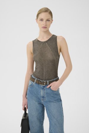 Vrouw in een mouwloze bruine gebreide top, blauwe jeans met een bruine riem, die een zwarte handtas vasthoudt, staande tegen een effen achtergrond.