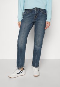 Jeans de mezclilla azul oscuro con corte recto, que cuentan con costuras blancas en contraste y un diseño de cinco bolsillos, combinados con zapatillas blancas.