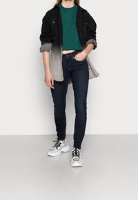 Chaqueta de mezclilla negra y gris sobre una camiseta recortada verde, combinada con jeans ajustados oscuros y zapatillas blancas con acentos azules.