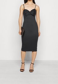 Svart figurnära klänning med en strukturerad rosettdetalj vid halsringningen, spaghettiband och knä-lång fåll. Matchad med svarta remsandaler.