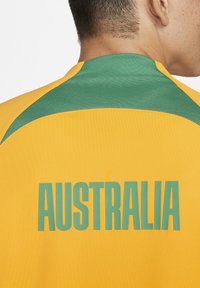 Gul sportsjersey med grønne detaljer på skuldrene. Store bogstaver på ryggen skriver "AUSTRALIEN" i grøn skrift. Glat stofstruktur.