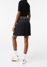 Černá plisovaná sukně s přiléhavým topem, zdobená bílým páskem s logem a kombinovaná s bílými teniskami a ponožkami. Hladká textury.