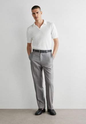 Mężczyzna ubrany w białą teksturowaną koszulkę polo z krótkim rękawem, szare eleganckie spodnie z czarnym paskiem i czarne eleganckie buty, stojący z rękami w kieszeniach.