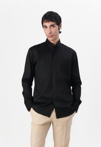 Μαύρο πουκάμισο με μακριά μανίκια και όρθιο γιακά, με λεία υφή και ραμμένο στυλ. Συνδυασμένο με μπεζ ραμμένα παντελόνια.