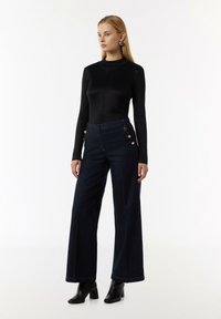 Czarny top z długim rękawem, dopasowany, w połączeniu z wysokim stanem, szerokimi spodniami z ciemnego denimu z dekoracyjnymi złotymi guzikami po bokach.