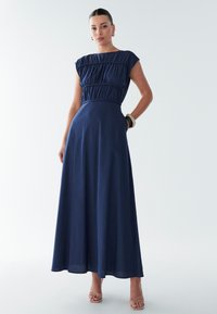 Tamsiai mėlyna maksi suknelė su klostuotu liemeniu, trumpomis rankovėmis ir ryškiai apibrėžta liemeniu. Pagaminta iš lengvos medžiagos, turi kišenes ir laisvai krentančią sijoną.