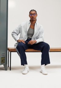 Model nosi prugastu majicu s dugim rukavima, sivu majicu bez rukava, mornarskoplave trenirke s bijelim prugama i bijele tenisice, sjedi na klupi.