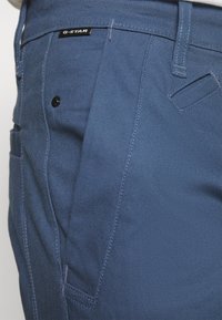 Blue G-Star pants with a textured fabric, featuring side pockets, a black button, and a branded label near the waistband.