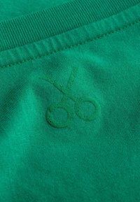 Groene stof met een zachte textuur heeft een reliëfschaarlogo. Het ribgebreide taillebanddetail aan de bovenkant past goed bij het gladde oppervlak.