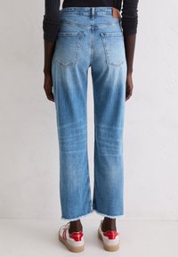 Personne portant un jean crop bleu clair avec des ourlets effilochés et des baskets blanches avec des accents rouges, se tenant contre un mur blanc.