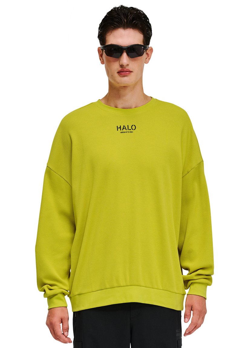 Limegrön oversize sweatshirt med ribbad struktur, rund halsringning och svart logotypdetalj. Modellen bär mörka solglasögon och svarta byxor.