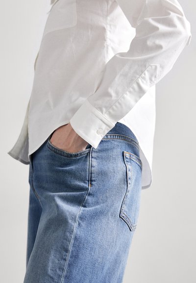 Cămașă albă din bumbac cu manșete butonate, asortată cu blugi din denim albastru deschis, având un buzunar la spate și o textură ușor estompată.