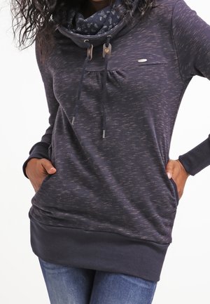 Kobieta ubrana w ciemny, długi sweter z kapturem, z troczkami, z rękami w kieszeniach, w połączeniu z niebieskimi dżinsami.