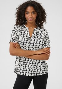 Blouse à manches courtes avec un motif abstrait noir et blanc, un col en V et un tissu doux et léger. Détails incluent des manches roulées.