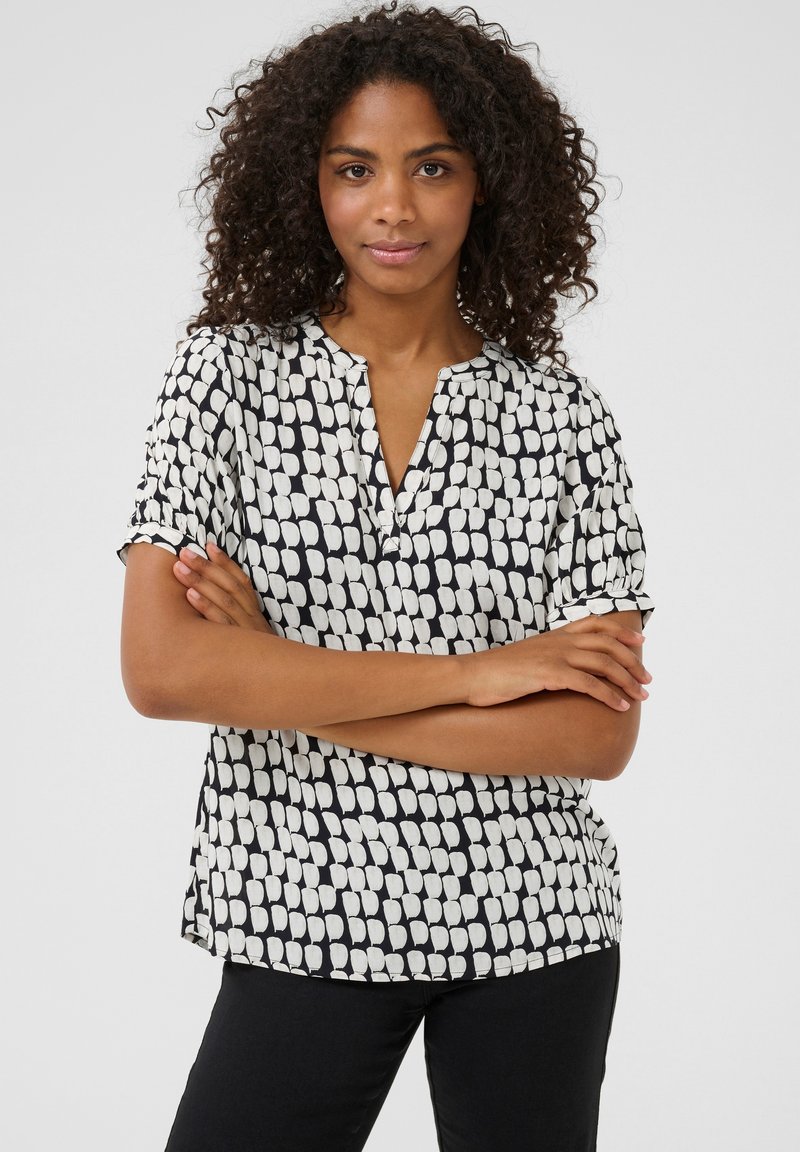 Blouse à manches courtes avec un motif abstrait noir et blanc, un col en V et un tissu doux et léger. Détails incluent des manches roulées.