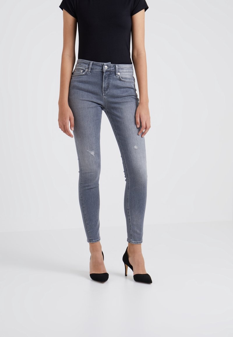 Jeans skinny de denim azul claro con detalles desgastados, diseño de talle alto y estilo de cinco bolsillos, combinados con tacones de aguja negros.