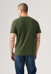 Bărbat întors, purtând un tricou verde măsliniu și blugi, cu tatuaje pe ambele brațe și o brățară argintie la încheietura stângă.