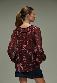 Gennemsigtig, maroon bluse med samlet halsudskæring, lange pufærmer og lyserøde mønstre af drikkevarer og sløjfer. Parret med en denimnederdel.