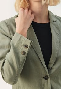 Zelený ľanový blazer s výstrihom, dvoma gombíkmi na manžetách a voľným strihom, kombinovaný s čiernym tričkom pod ním.