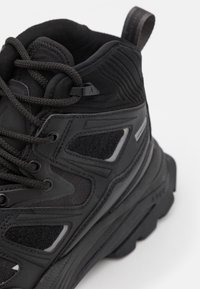 Černá turistická obuv s texturovaným nylonovým svrškem, gumovými doplňky, zesíleným šněrovacím systémem a pevnou podrážkou se vzorkem pro lepší trakci.