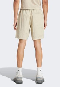 Pantaloni corti in cotone beige con vita elastica, una sola tasca posteriore e una vestibilità rilassata, abbinati a sneaker grigie e calzettoni bianchi.