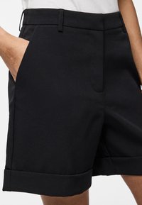 Calções pretos ajustados com corte reto, bolsos laterais, textura suave e bainhas viradas. Feitos de um tecido leve.
