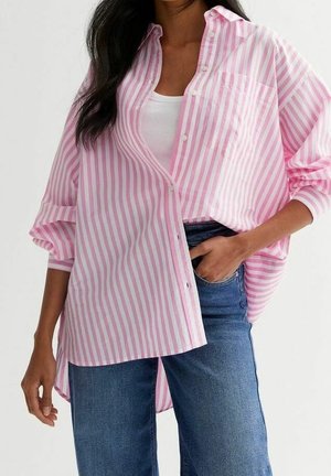 Žena nosící přehnaně velkou růžovo-bílou proužkovanou košili přes bílý top bez rukávů, v kombinaci s modrými džínami, s jednou rukou v kapse.